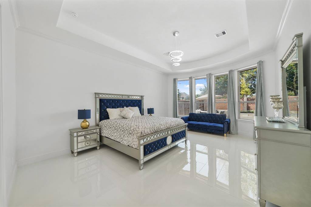 The extra large master bedroom features a cozy seating area, a wall of windows that offer a serene view of the pool, a tray ceiling that adds architectural elegance, polished tile floors, and a beautiful chandelier, creating a luxurious. and tranquil sanctuary.