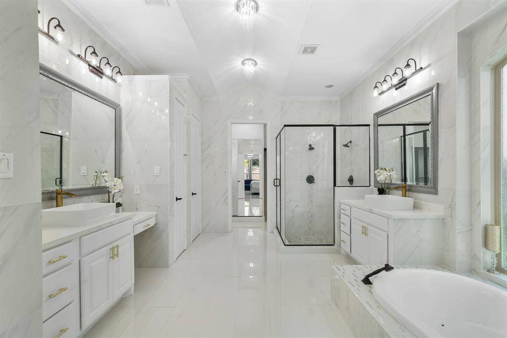 The master bathroom is a luxurious retreat, covered wall to wall in gleaming polished tiles, featuring stunning chandeliers that cast a warm glow separate his and her vanities, a soothing hot tub jetted spa, a spacious standalone shower with 2 shower-heads, and a private restroom, creating a perfect blend of elegance and comfort.