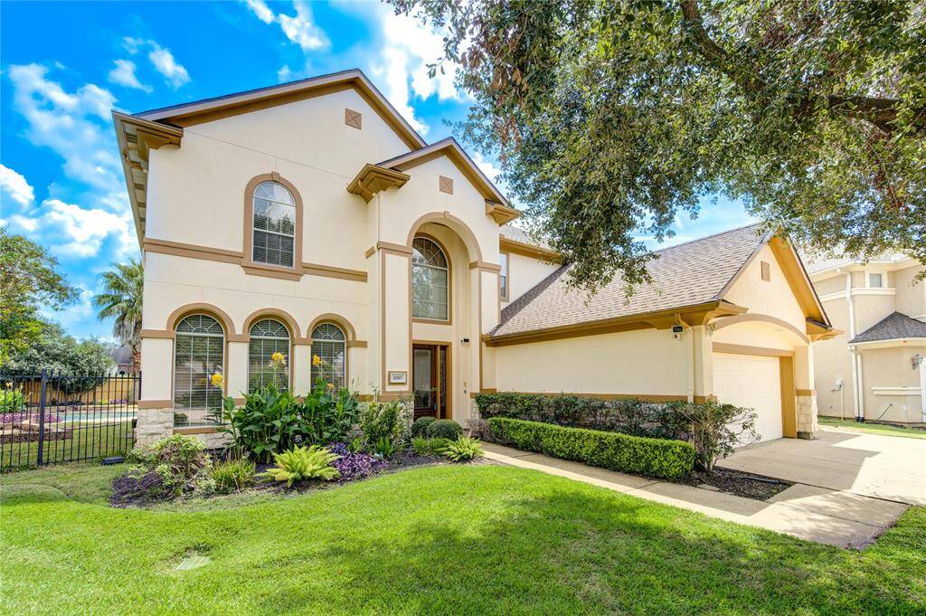This is a two-story home featuring a beige stucco exterior with stone accents, arched windows, and a side-entry two-car garage. The property is well-manicured with mature landscaping and a green lawn, situated under a clear blue sky.