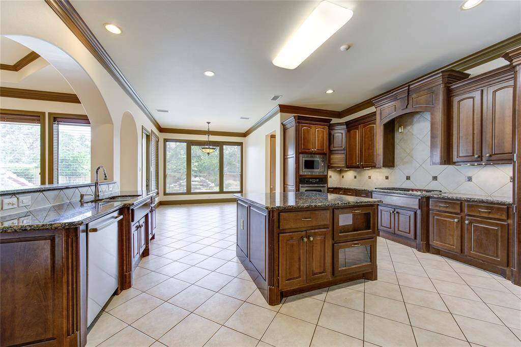 This spacious kitchen features rich wooden cabinetry, granite countertops, and a modern stainless steel oven and cooktop. The open layout offers plenty of natural light from large windows, with a view towards the dining area. Perfect for both cooking and entertaining.