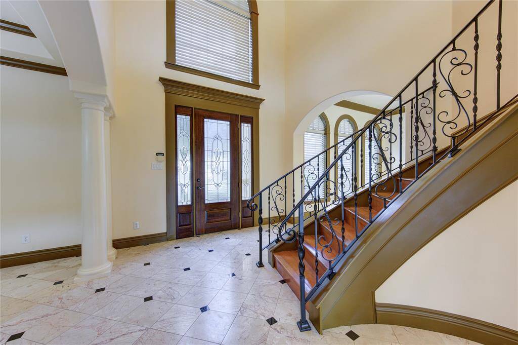 This is an elegant home office or formal sitting area, featuring large arched windows with blinds, a matching bookshelf, and a stylish area rug (virtually staged) on polished marble flooring. The room is finished with crown molding, warm lighting, and modern art pieces, creating a luxurious workspace.