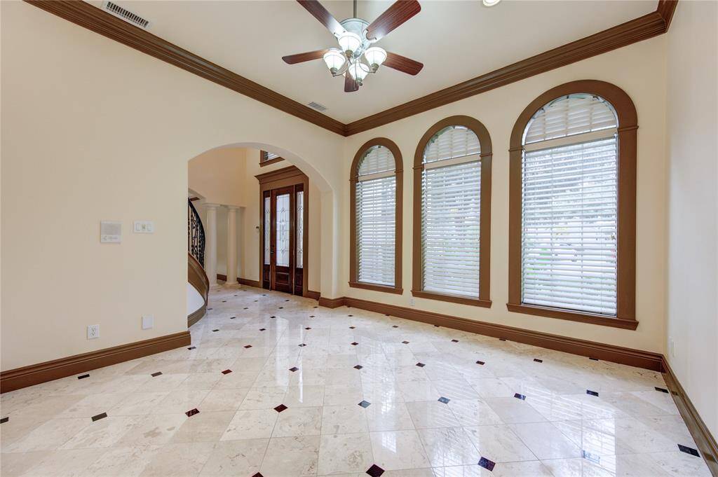 This photo showcases an elegant dining area with a large wooden table and plush green chairs (virtually staged), complemented by a classic chandelier. The room features a coffered ceiling with decorative moldings and an open archway leading to a grand staircase, providing a glimpse into the home's stately architecture and luxurious details.