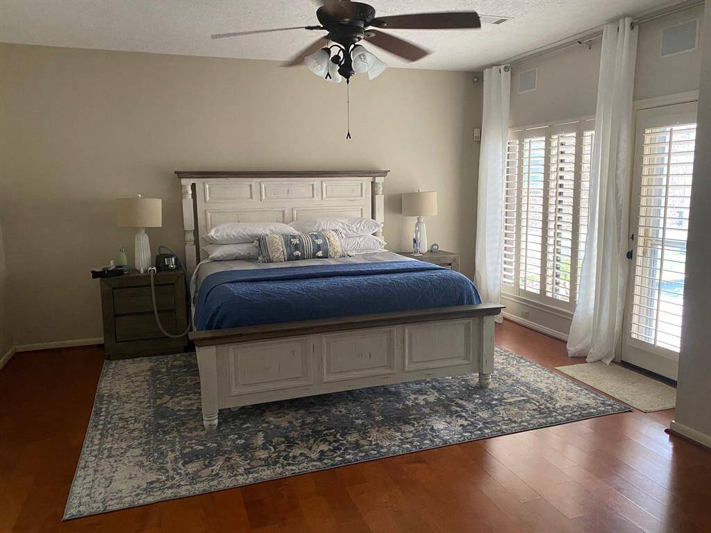 Spacious primary bedroom featuring elegant hardwood floors, abundant natural light from large windows with plantation shutters, and a charming ceiling fan for added comfort.