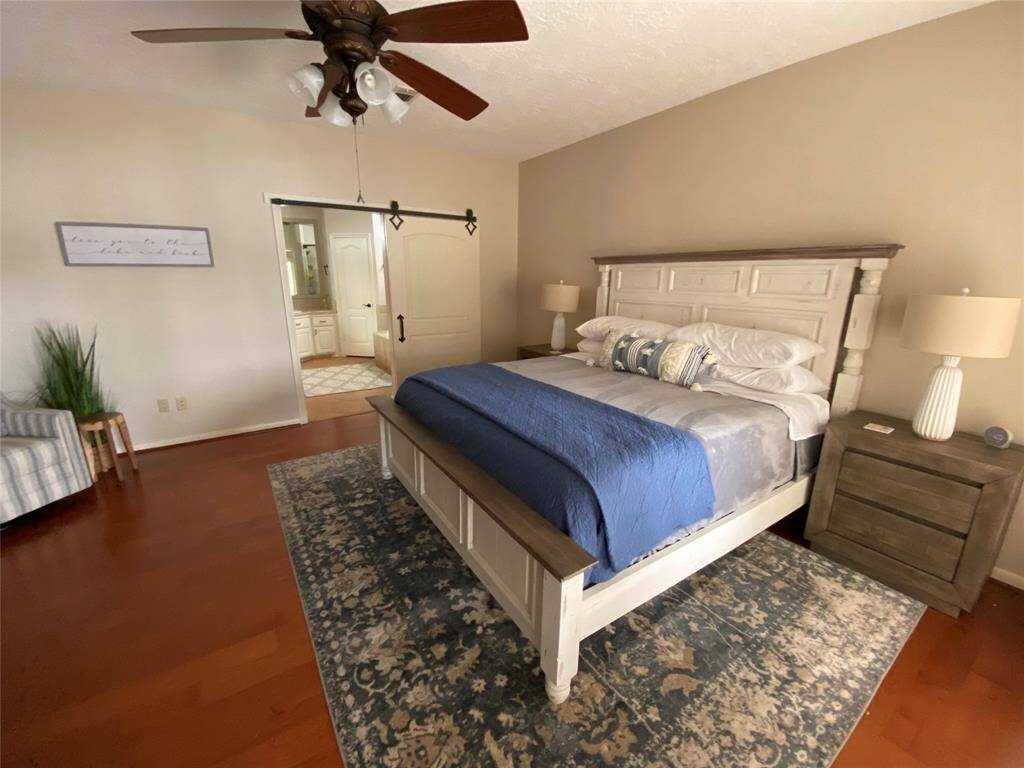 This photo features a spacious bedroom with hardwood floors, a ceiling fan, and a large bed with a stylish headboard. The room is tastefully decorated with bedside tables, lamps, and a decorative rug. A sliding barn door leads to an en-suite bathroom, enhancing the room's modern and cozy feel.