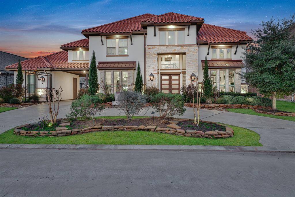 The stunning two-story home with a Mediterranean design, featuring a red-tiled roof and stone accents. The property is beautifully landscaped with manicured shrubs and trees, and a spacious stamped driveway leading to a gated entry. The exterior lighting enhances the home's elegance at dusk.