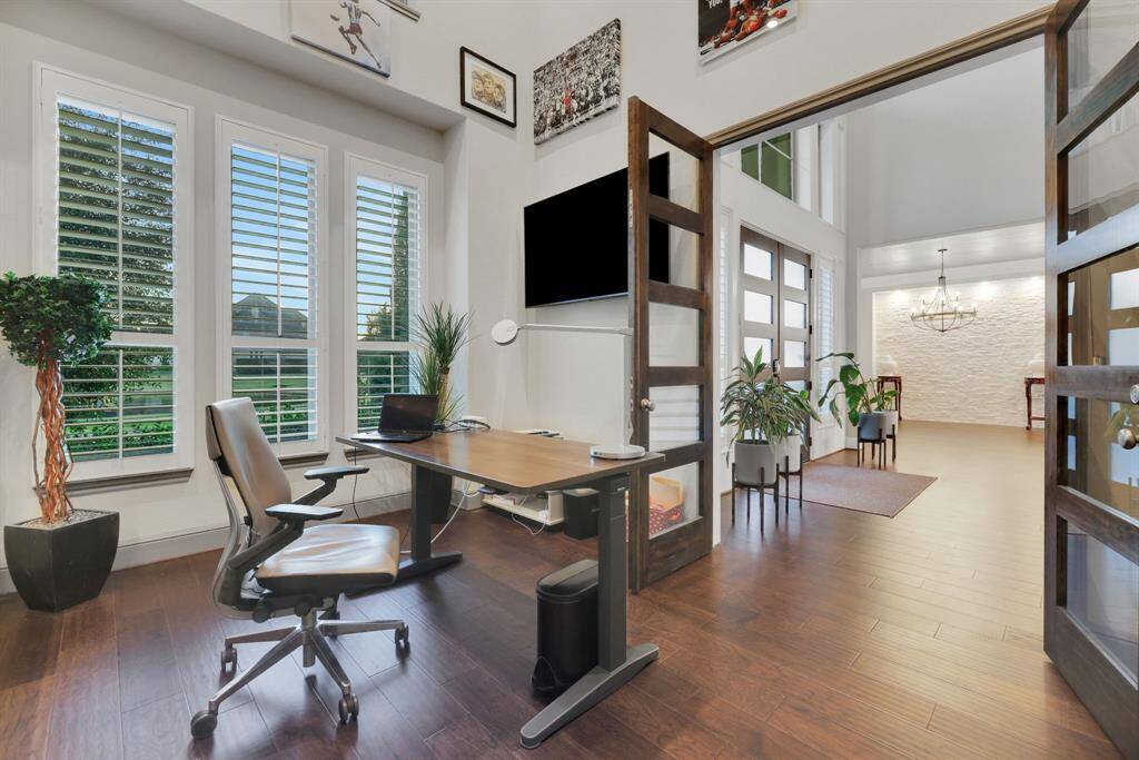 A modern home office with hardwood floors, large windows with plantation shutters, and ample natural light. It features high ceilings, custom cabinetry and wood-lay ceilings.
