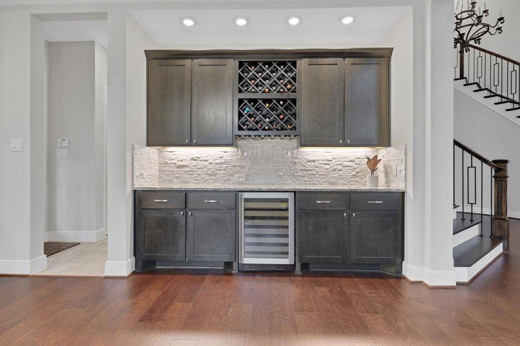 A stylish home dry bar area with dark wood cabinets, a built-in wine rack, and a sleek wine cooler. The backsplash features mosaic stone tiles, and recessed lighting enhances the elegant design. Adjacent to stairs, it offers a modern, inviting space ideal for entertaining.