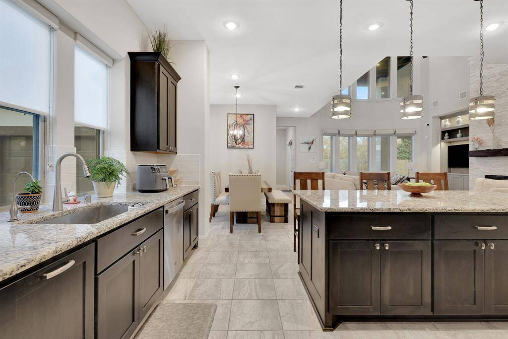 This modern kitchen and dining area features sleek granite countertops, dark cabinetry, and stainless steel appliances. It opens to a bright living space with large windows and contemporary lighting, creating an inviting atmosphere ideal for entertaining.