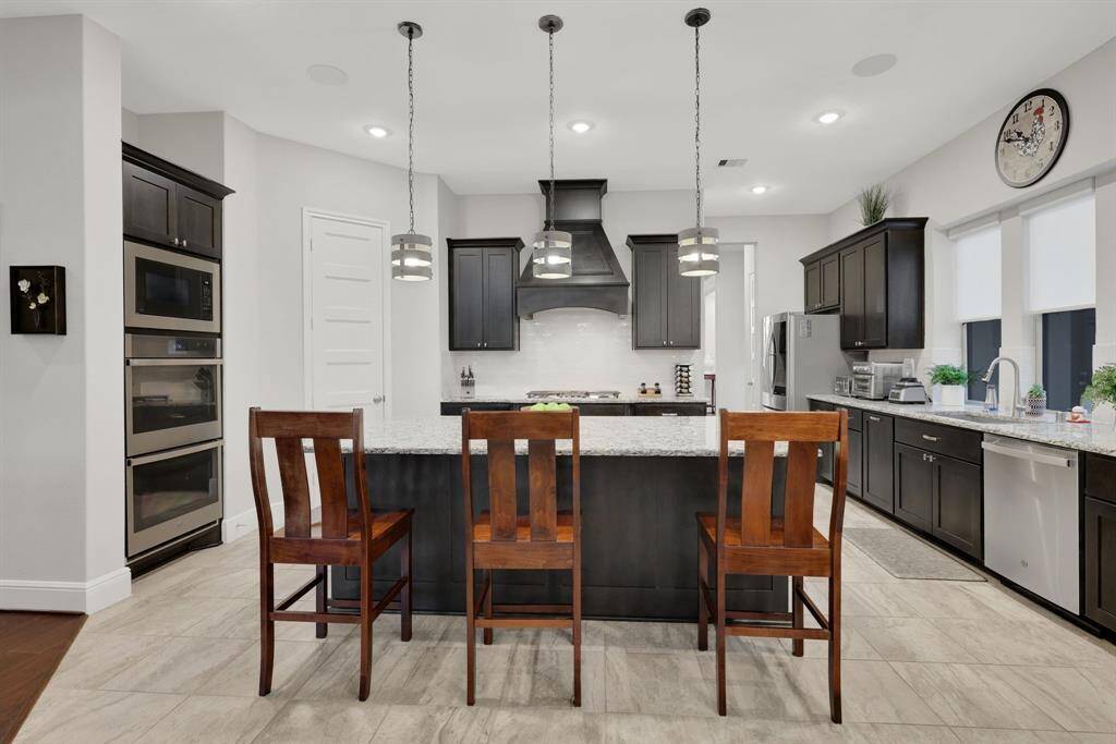 This modern kitchen features sleek dark cabinetry, a large granite island with seating, stainless steel appliances, and stylish pendant lighting. The space is bright and welcoming, ideal for cooking and entertaining.