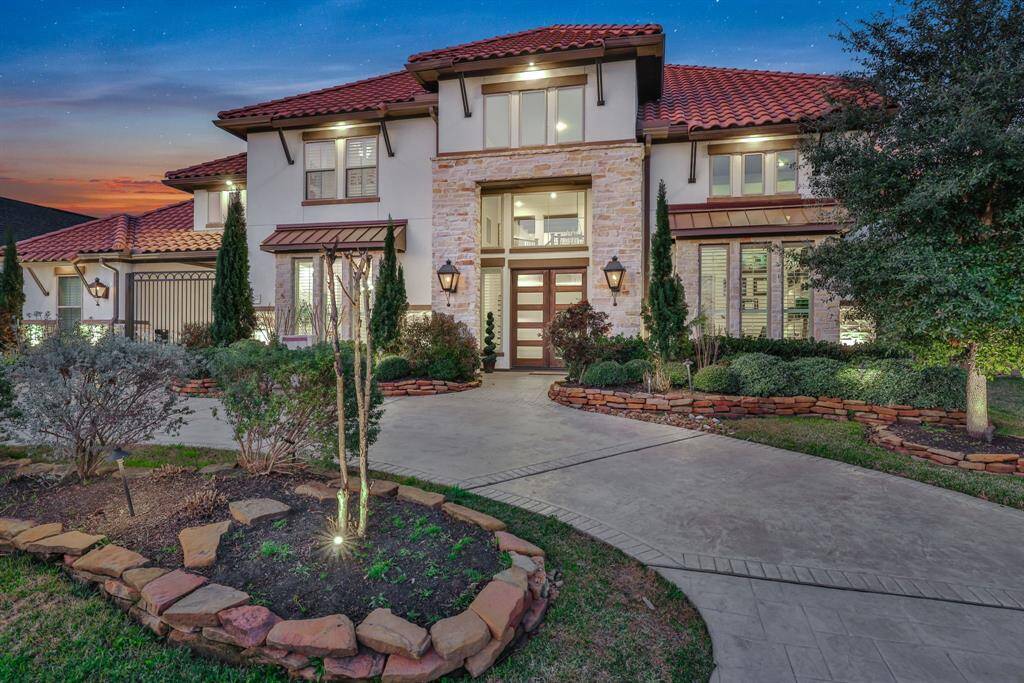 The stunning two-story home features a modern design with a red-tiled roof, grand entrance and stamped concrete driveway. It is beautifully landscaped with stone-bordered gardens, and the well-lit exterior enhances its curb appeal.