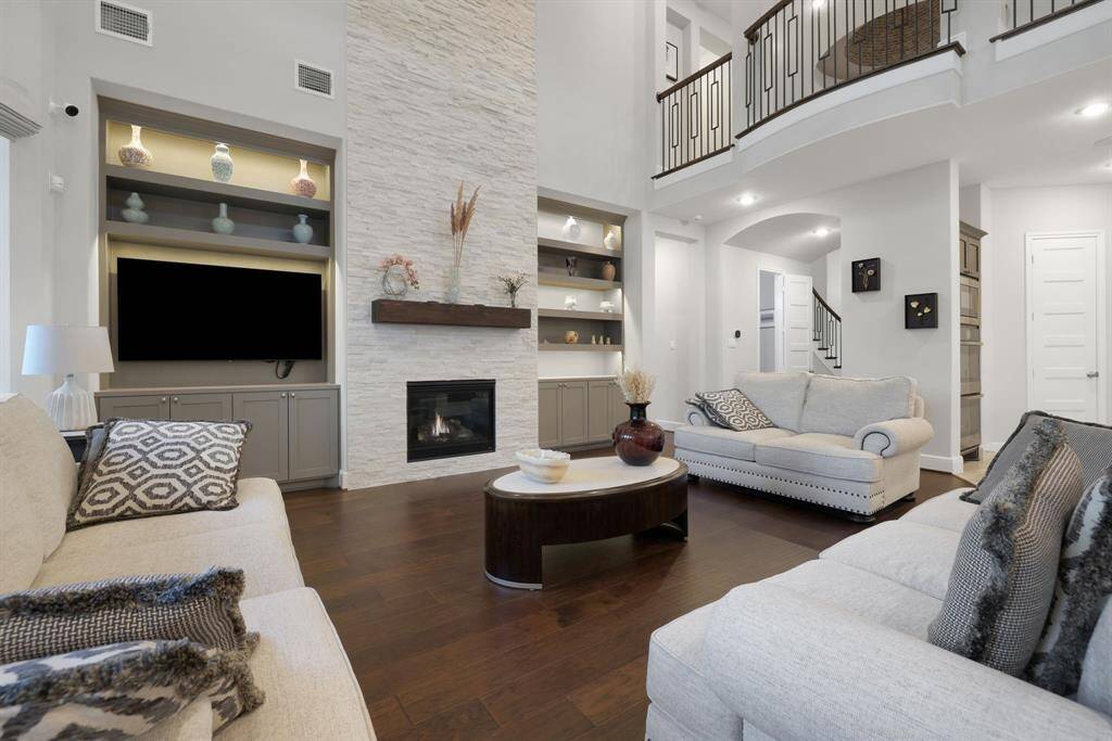 This spacious living room features high ceilings, a cozy fireplace, and elegant built-in shelves. The open design includes a modern color palette with neutral tones, hardwood floors, and a stylish staircase leading to an upper level. Ideal for comfortable living and entertaining.