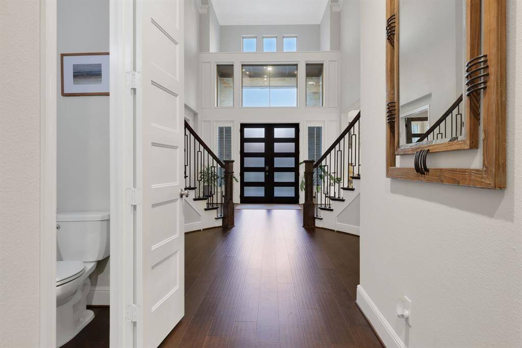 This photo showcases a modern, spacious entryway with high ceilings and abundant natural light. It features elegant dark wood flooring, a stylish front door with frosted glass panels, and a grand staircase with upgraded wrought iron railings. A sleek powder room is conveniently located to the side.