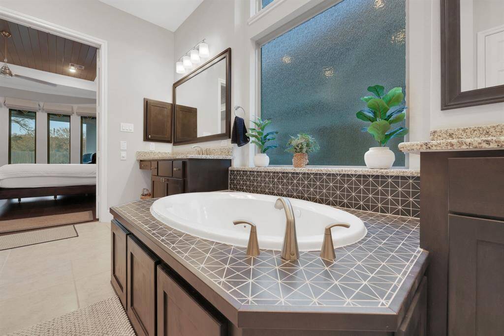 The modern bathroom with a large soaking tub, elegant tile surround, and dual sinks with granite countertops. The space is brightened by a large frosted window.