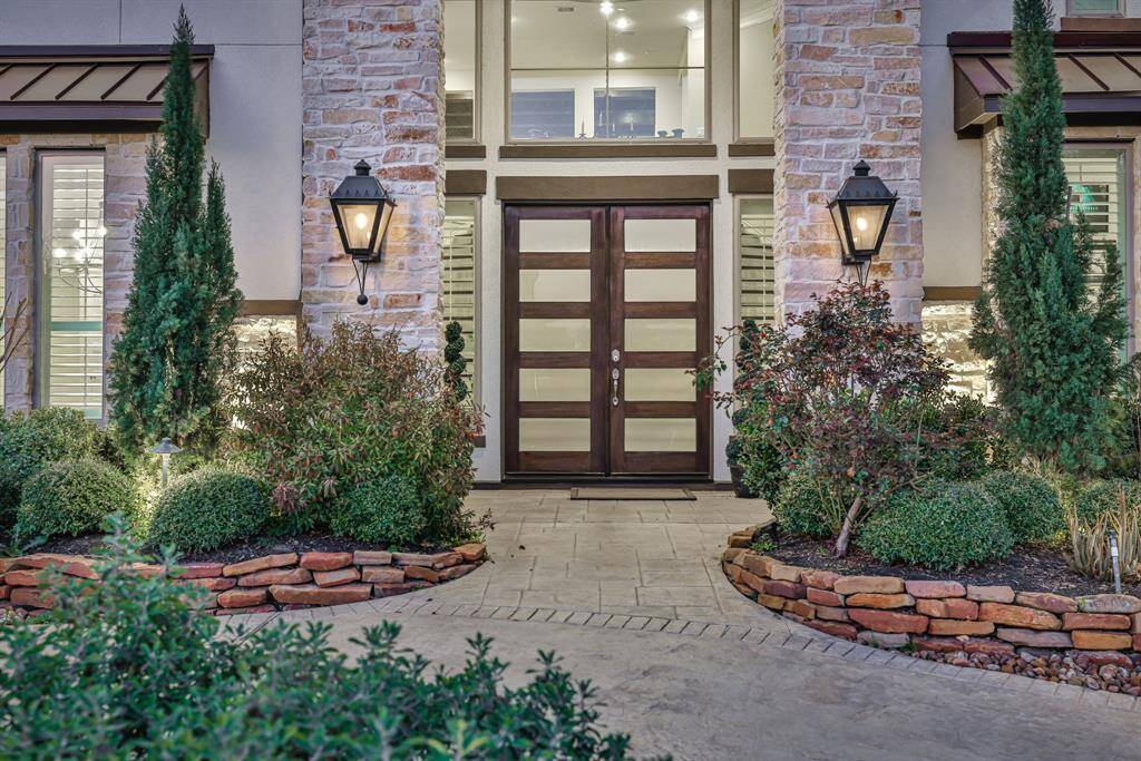 The stylish home entrance with a modern glass-panel door framed by stone accents and elegant lantern lighting. The landscaped pathway features neatly arranged shrubs and small trees, creating an inviting and sophisticated curb appeal.