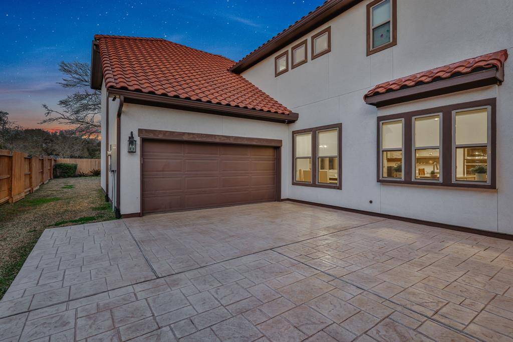 The modern home with a three-car tandem garage, featuring a tiled roof and a spacious paved driveway. The exterior has large, well-framed windows that hint at a bright interior, and the property is bordered by a wooden fence, offering privacy and a touch of nature.