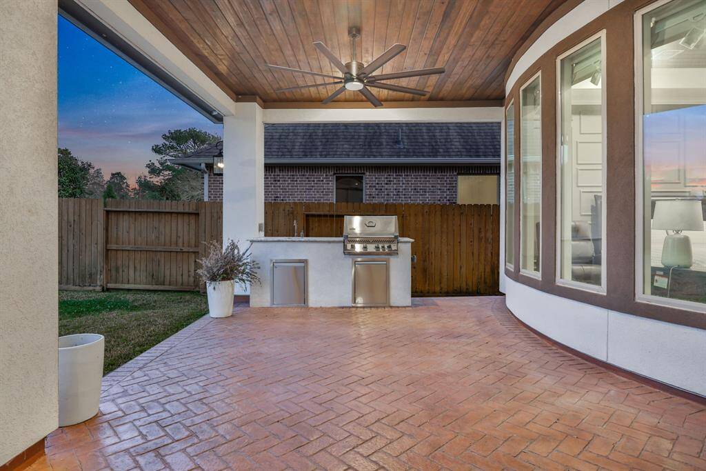 The stylish outdoor patio with a built-in grill, under a wooden ceiling with a fan. It features a fenced yard and large, curved windows, offering a perfect space for entertaining. Pre-wired for outdoor speakers.