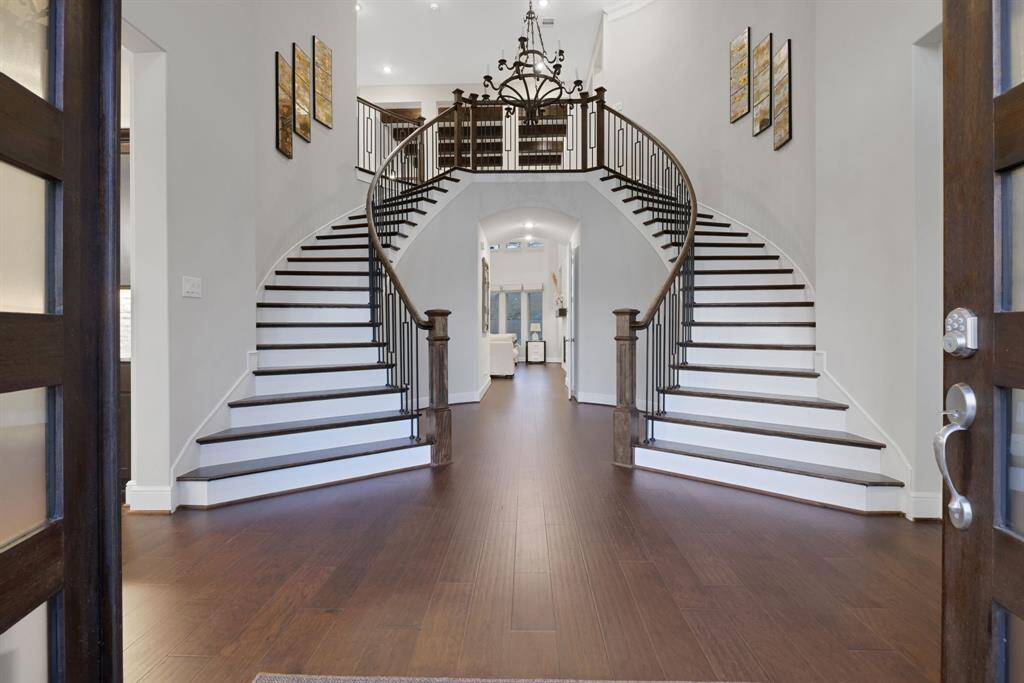 The grand foyer with a stunning double staircase, dark wood flooring, and elegant railings. The space is bright and welcoming, featuring a high ceiling and a central chandelier, creating a luxurious entrance.