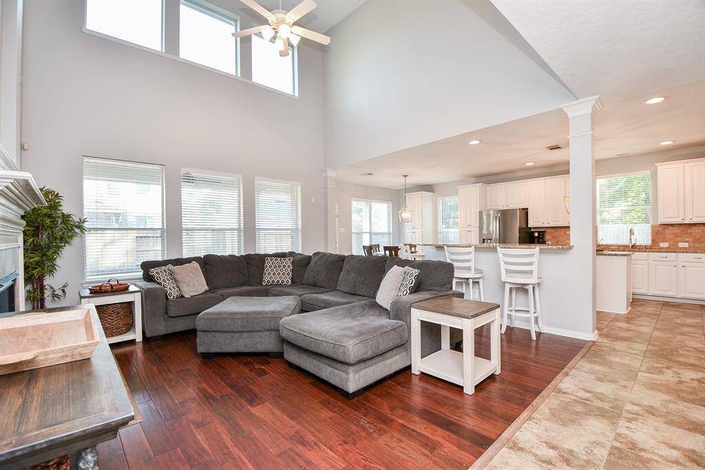 This spacious open-concept living area features high ceilings and abundant natural light. It includes a cozy sectional, hardwood flooring, and a seamless flow into a modern kitchen with a breakfast bar, perfect for entertaining.
