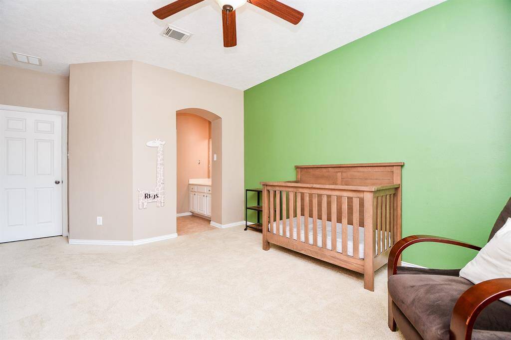 This cozy room features a soft beige carpet, a vibrant green accent wall, and a ceiling fan for comfort. It's furnished with a wooden crib and a cushioned chair, making it ideal for a nursery. An arched doorway leads to a small vanity area, adding convenience.