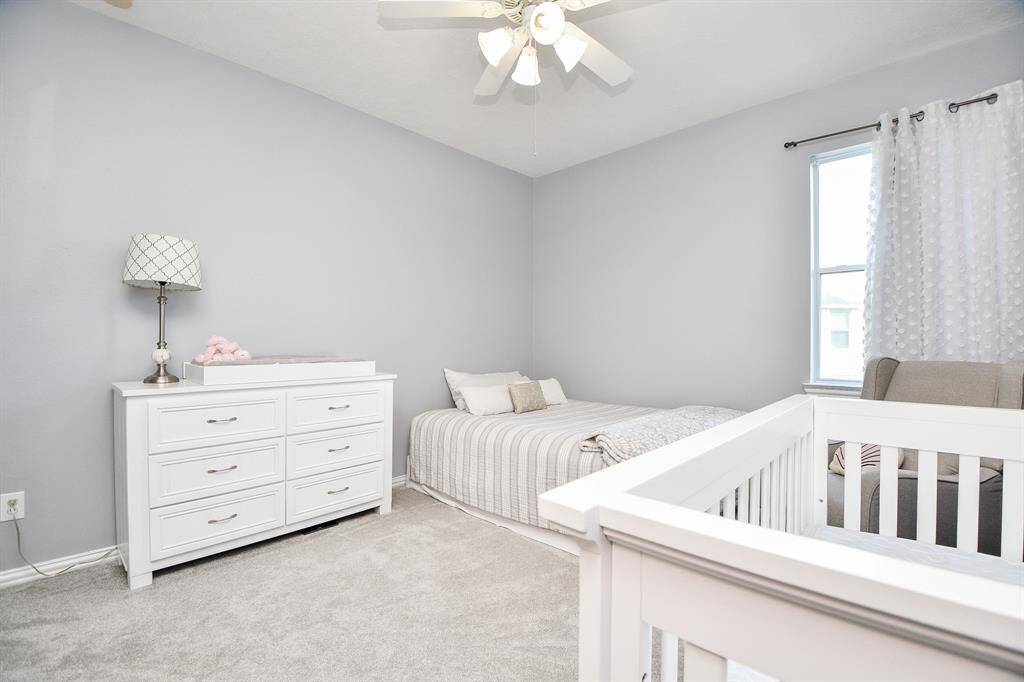 This cozy nursery features soft gray walls, a white dresser, a crib, and a comfortable bed. It has plush carpeting, a ceiling fan, and a window with sheer curtains, creating a bright and welcoming atmosphere. Ideal for a child's room or guest space.
