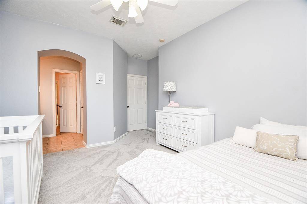 This bedroom features soft gray walls and carpet, creating a serene atmosphere. It includes a ceiling fan, a white dresser with a lamp, and a cozy bed. An arched doorway leads to another room, adding character to the space.