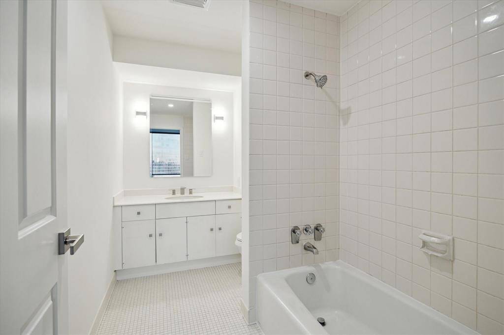 This bathroom features a clean, minimalist design with white tiles throughout. It includes a bathtub with a showerhead, a large mirror above a sleek vanity, and modern fixtures, creating a bright and airy space.