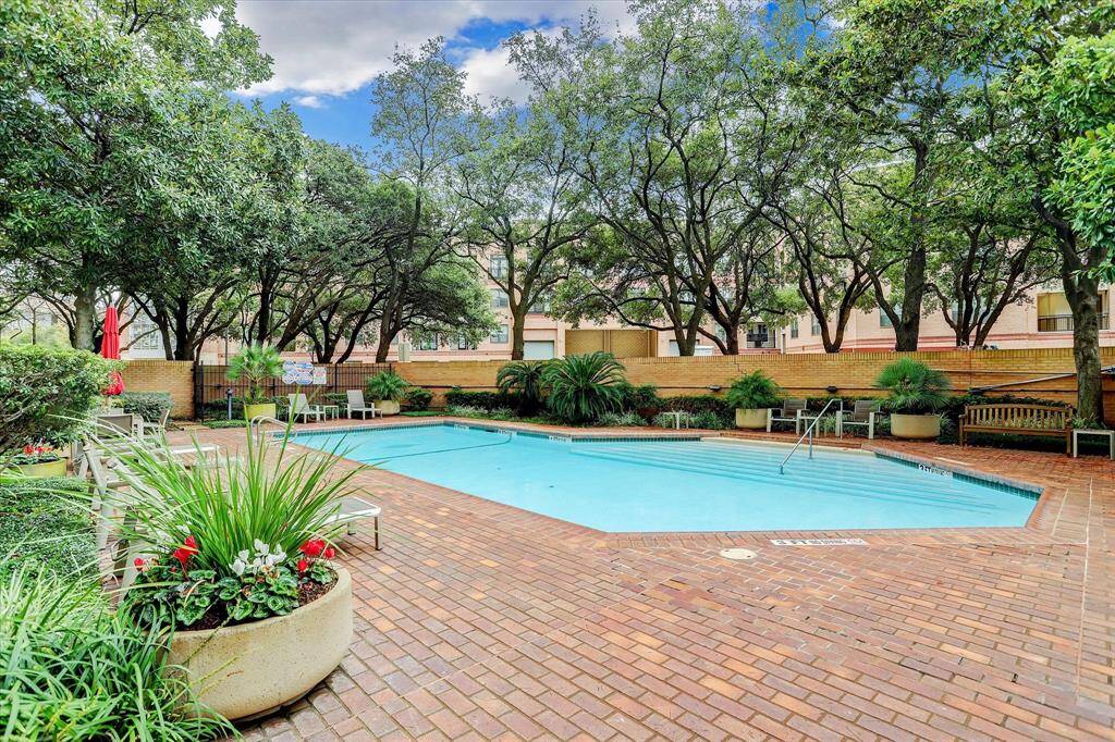 A serene outdoor area featuring a heated swimming pool surrounded by lush greenery and mature trees. The brick patio offers ample seating, creating a relaxing environment perfect for leisure and entertaining.