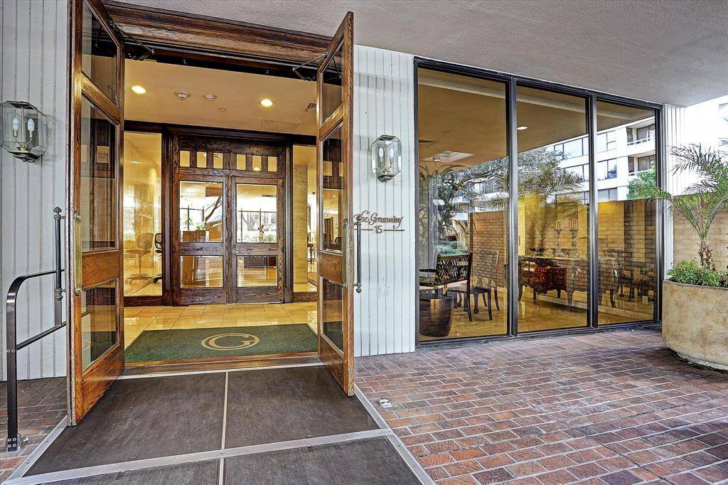 The inviting entrance of 15 Greenway features large wooden doors and expansive windows. The entrance is well-lit and welcoming, with a glimpse of a stylish lobby area inside. The exterior is accented with brick flooring and potted plants, creating a modern and elegant feel.