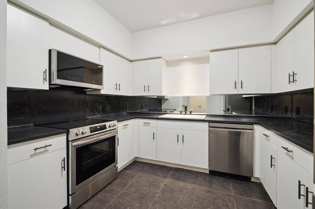 Modern kitchen with new farmhouse sink with mixed metal fixtures, sleek black countertops and backsplash, white cabinetry, new stainless steel appliances, and tiled flooring.