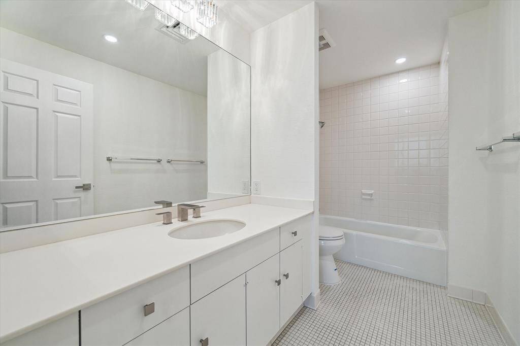 The secondary bathroom features a clean, modern design with a large mirror, sleek white cabinetry, and a tiled bathtub area. The space is well-lit with new contemporary fixtures and neutral decor.