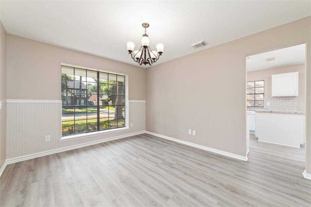 This light and bright room with modern gray luxury vinyl plank flooring and large windows that provide ample natural light. The space features neutral wall colors, wainscoting details, and a view into an elegantly appointed kitchen.