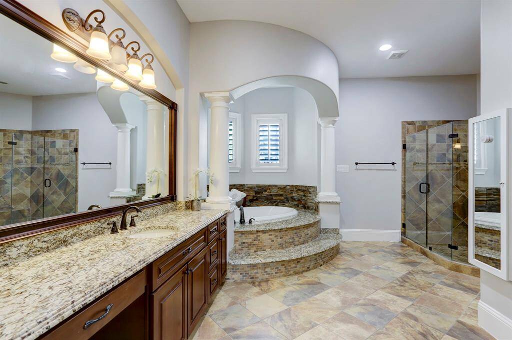 A double-sink vanity with rich wood cabinetry and granite countertops offers ample storage and a refined focal point, while the large mirror and stylish lighting enhance the sense of openness. The neutral palette and thoughtful architectural details, such as the curved archway and column accents, contribute to an inviting, sophisticated ambiance—ideal for those seeking a tranquil retreat in their own home.