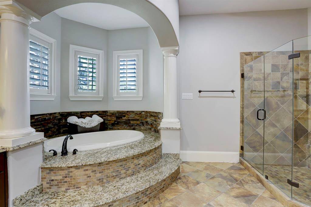 This spacious, spa-inspired primary bathroom exudes luxury with its grand arched entrance and stately columns framing an elevated soaking tub. The warm, neutral tile flooring extends seamlessly to the tub surround and the generously sized, glass-enclosed walk-in shower, creating a cohesive and elegant look.