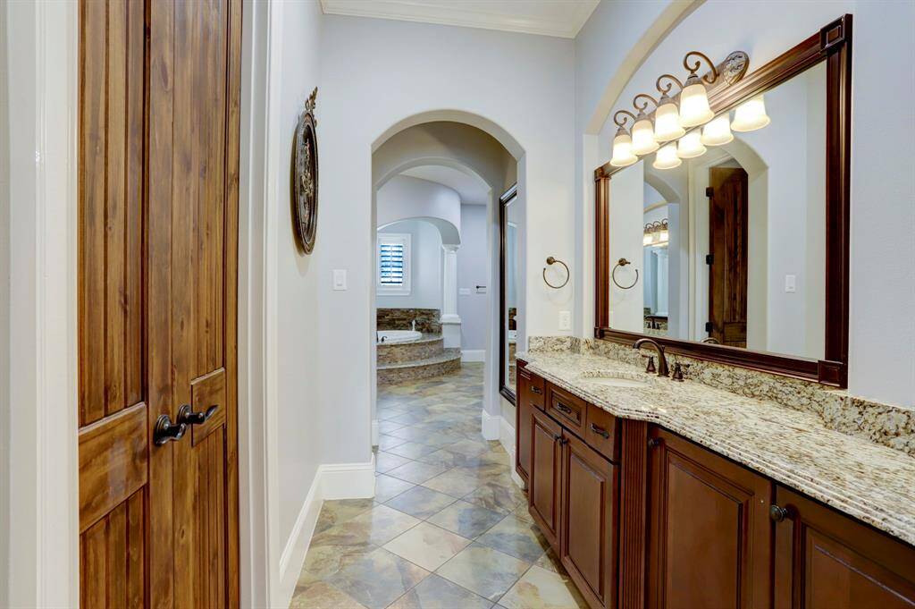 This photo showcases one of two separate double-sink vanities in the primary bath, offering abundant counter space and a rich, warm aesthetic. Granite countertops, elegant arches, and wood doors create a sophisticated yet welcoming environment. The neutral tile flooring flows seamlessly through the space, leading to the luxurious, elevated tub area beyond.
