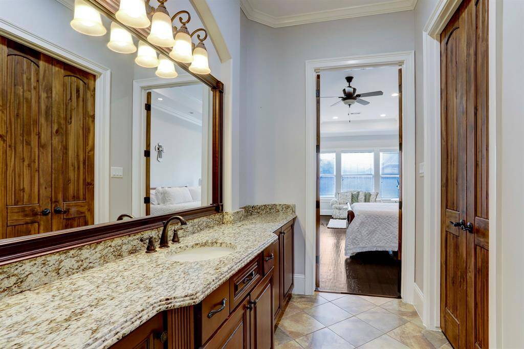 This photo offers a glimpse into the seamless flow between the primary bath and primary bedroom, highlighting the warm elegance of the wood doors and rich granite vanity. Ample counter space and under-sink storage make daily routines convenient, while the neutral tile flooring ties the spaces together.