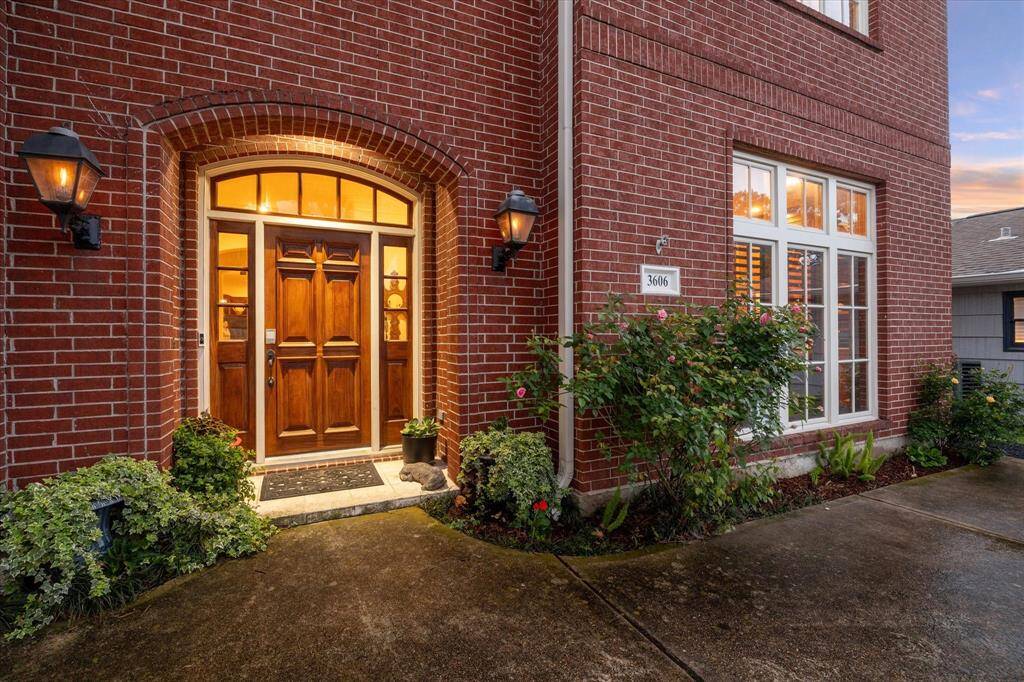 Inviting arched entryway with a mahogany front door with elegant gas lighting, and large windows that allow for ample natural light. Landscaping includes lush greenery and flowering plants, enhancing curb appeal.