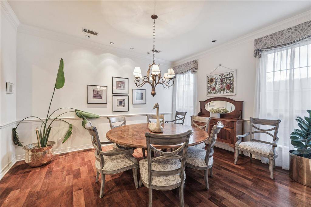 This is an elegant dining area featuring hardwood floors, a classic wooden dining table with matching chairs, and a traditional chandelier. The room is well-lit by natural light from large windows with stylish draperies and is adorned with wall art and plants, creating a warm and inviting space.