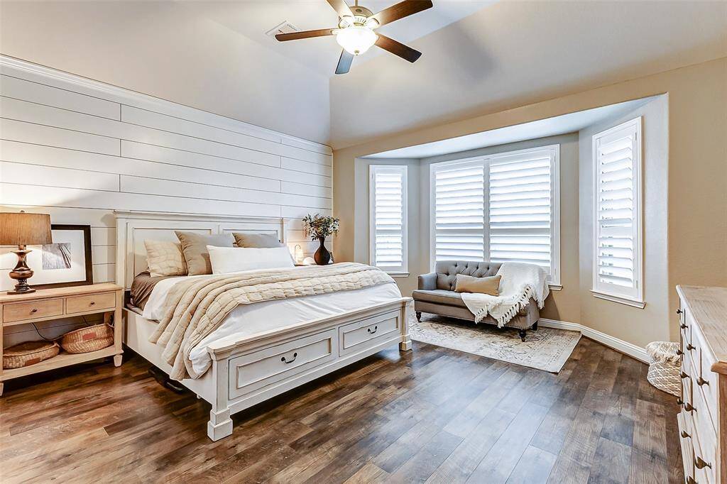 Welcome to your beautiful primary suite! Highlights include a stylish shiplap accent wall, a raised ceiling, and a stunning bay window with plantation shutters, providing the perfect spot for a cozy seating area.