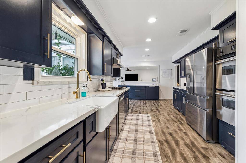 The kitchen features cabinets with gold hardware, white countertops, and a farmhouse sink. It offers stainless steel appliances, including double ovens. The space is well-lit with recessed lighting and has a modern, open feel with wood flooring.