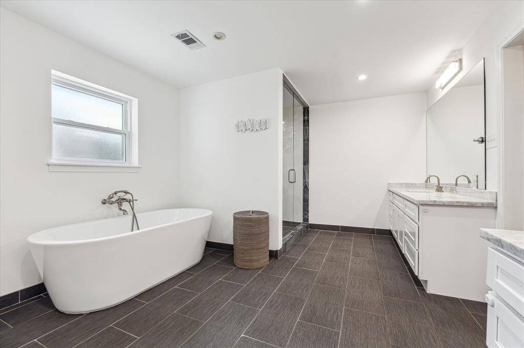 The modern primary bathroom features a freestanding tub, a spacious glass-enclosed shower, and a double vanity with ample storage. The room is bright with a window and contemporary fixtures, complemented by dark tile flooring.