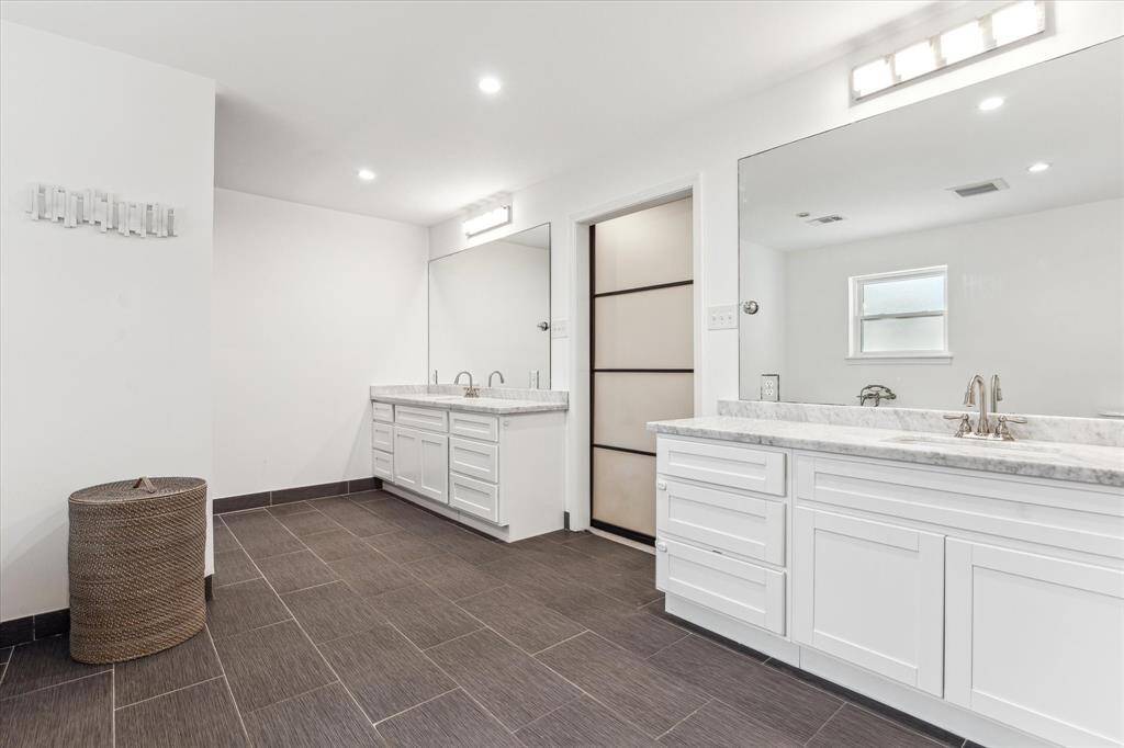 The primary bathroom features a spacious and modern design with dual sinks, elegant white cabinetry, and sleek dark floor tiles. Ample lighting and a large mirror enhance the bright, clean aesthetic.