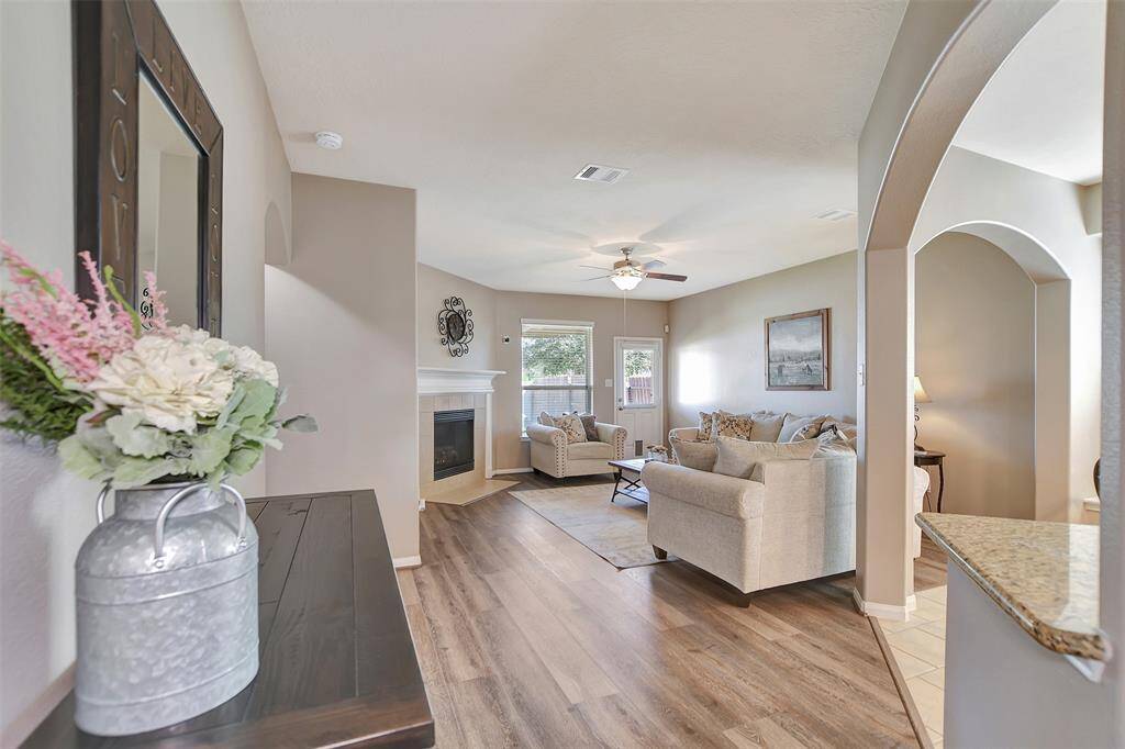Bright, open living room with vinyl plank flooring, cozy fireplace, and neutral tones. Features a ceiling fan, large windows for natural light, and an adjacent arched entryway leading to other areas. Perfect for comfortable living and entertaining.