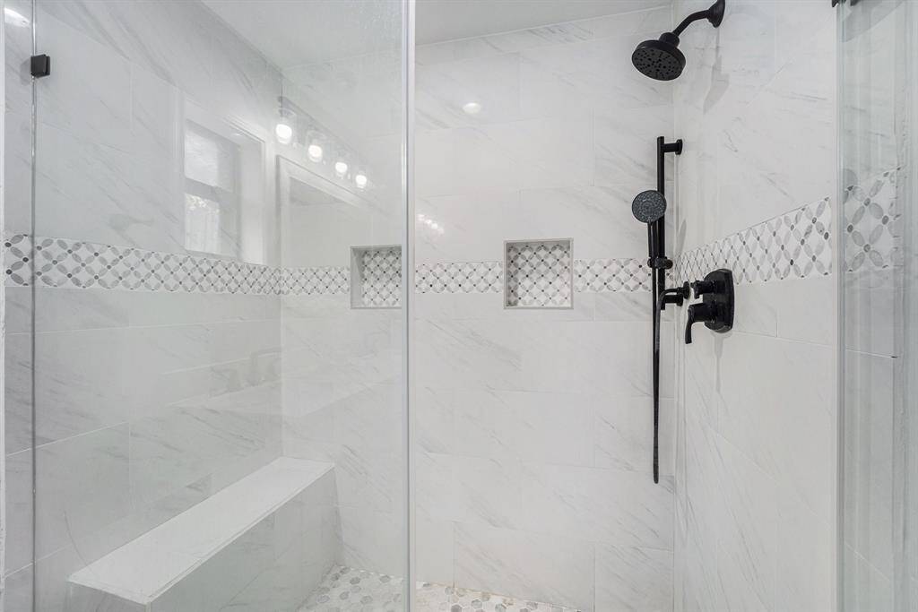 Framless shower with elegant white marble tiles and a decorative mosaic strip. It features a bench, dual shower heads, and built-in niches for storage.