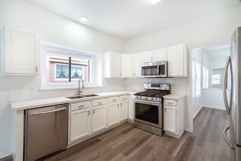 Bright and modern kitchen featuring white cabinetry, stainless steel appliances, and hardwood flooring. A large window provides natural light, and there's ample counter space for cooking.