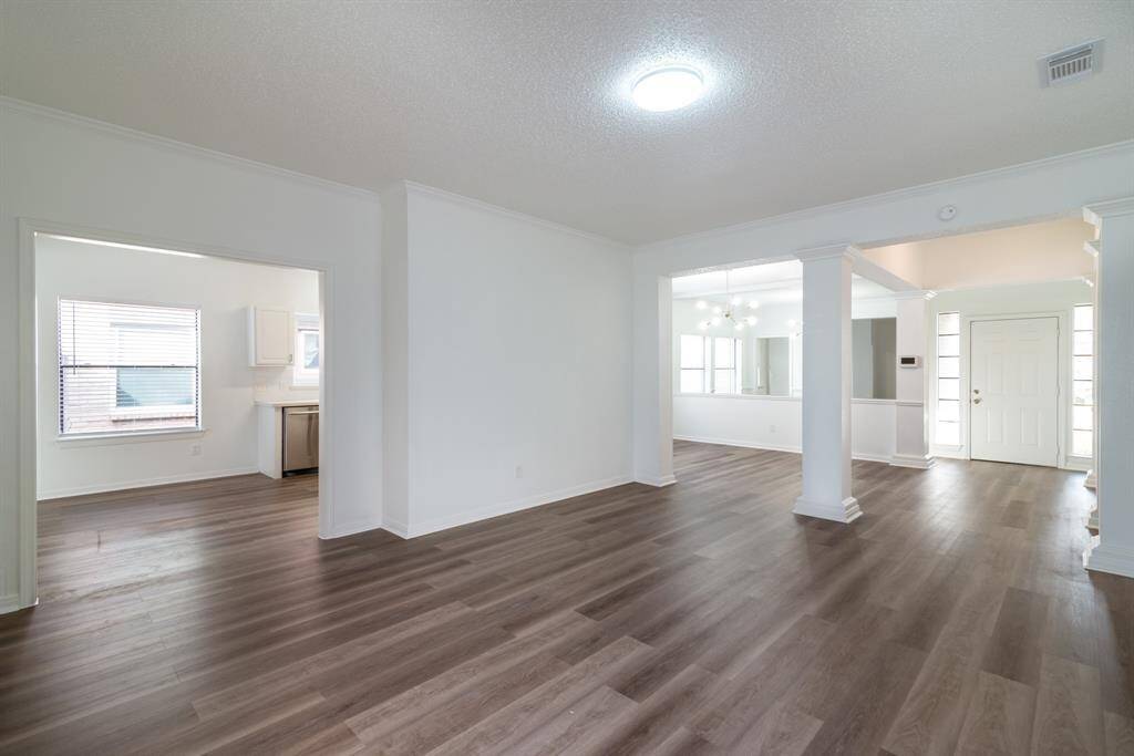 Spacious open-plan living area with modern flooring and ample natural light. It features a view into a bright kitchen, a dining space with stylish columns, and a welcoming entryway.