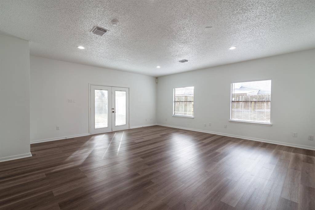 This spacious, bright living area features sleek wood flooring, ample natural light from multiple windows, and French doors leading outside. Ideal for comfortable living and entertaining.