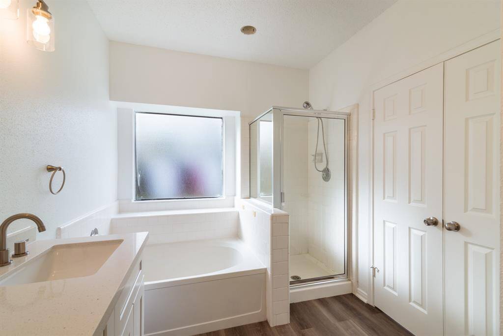 This bathroom features a modern design with a white color scheme. It includes a spacious bathtub, a separate glass-enclosed shower, a sleek vanity with a large mirror, and ample natural light from a frosted window. The room has wood-style flooring and double-door storage.