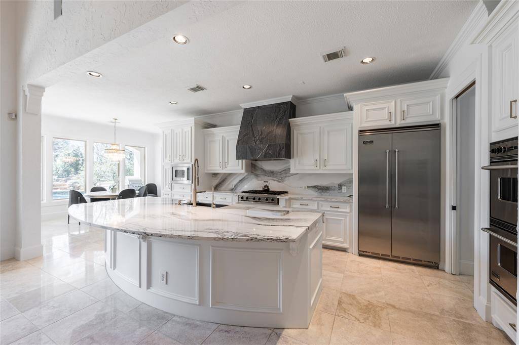 This kitchen features a spacious, modern design with a large quartz-topped island, stainless steel appliances, and ample white cabinetry. Natural light floods in through large windows, creating a bright and inviting atmosphere.