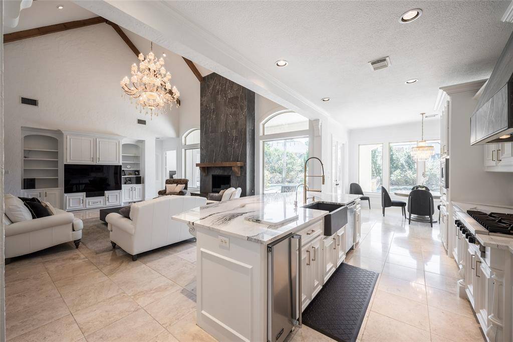 This photo showcases a spacious open-concept living area with high ceilings and elegant finishes. The kitchen features a large island with a farmhouse sink, quartz countertops, and modern appliances. The living room includes a cozy fireplace and built-in shelving, all illuminated by a stunning chandelier. Large windows provide plenty of natural light and a view of the outdoors.