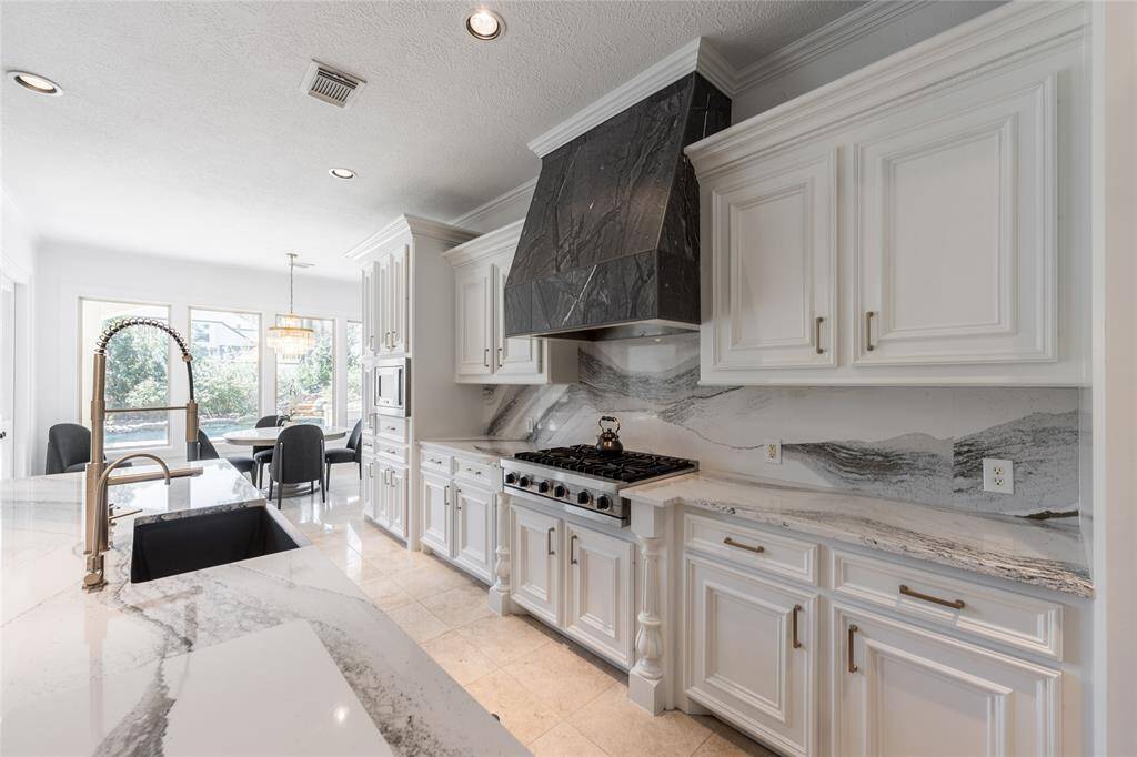 This kitchen features elegant white cabinetry, a sleek quartz countertop, and a stylish quartz backsplash. It includes a modern gas stove with a dark, striking range hood. Natural light floods in from large windows, illuminating the spacious area with a built-in dining space for a cozy ambiance.